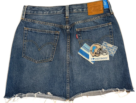 A pair of blue denim shorts with raw, frayed hems, laid flat on a white background. The shorts feature classic five-pocket styling, a brown leather patch on the right side, and a small blue Levi's brand tab on the left side.