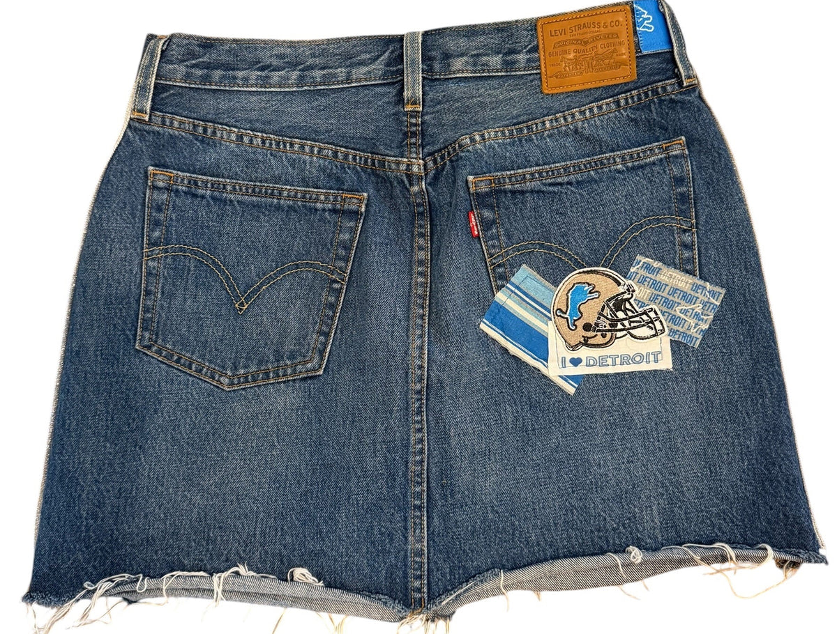 A pair of blue denim shorts with raw, frayed hems, laid flat on a white background. The shorts feature classic five-pocket styling, a brown leather patch on the right side, and a small blue Levi's brand tab on the left side.