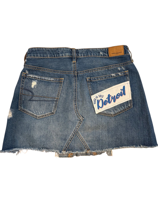 Back view of a distressed blue denim mini skirt with a frayed hem, featuring a 'Detroit' patch on the right pocket and a brown leather brand tab at the waistband.