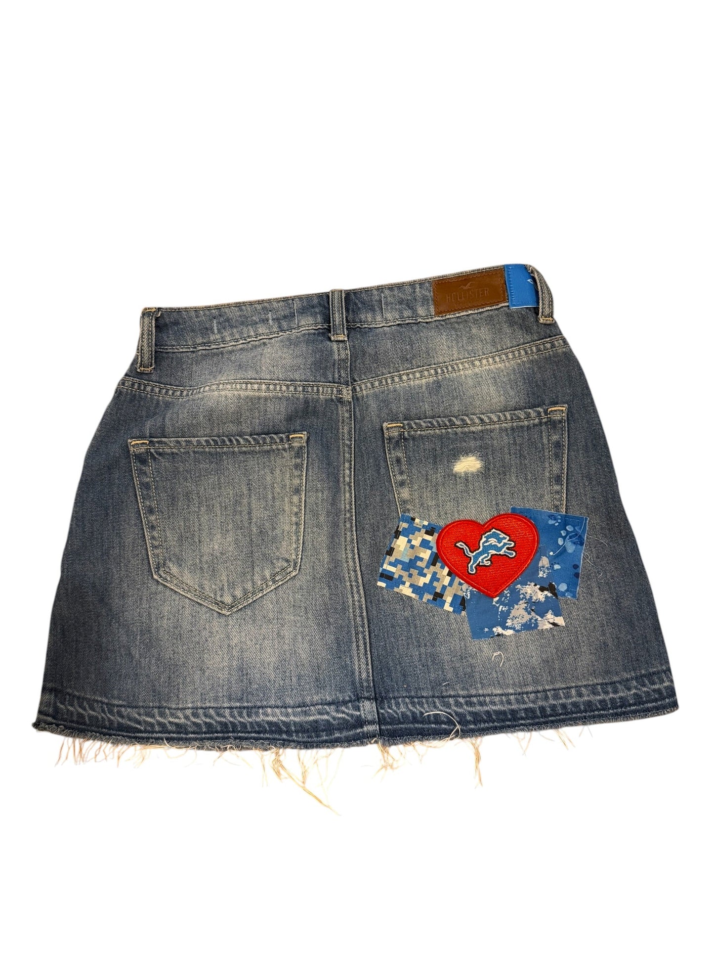 A denim skirt with a raw, frayed hem and distressed details, featuring a colorful patch on the back pocket with a red heart and blue patterned fabric.