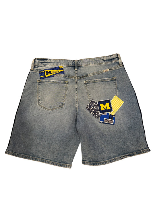 Light wash denim shorts shown from the back, featuring a blue and yellow Michigan Wolverines patch on the right back pocket and a smaller matching patch on the left back pocket.