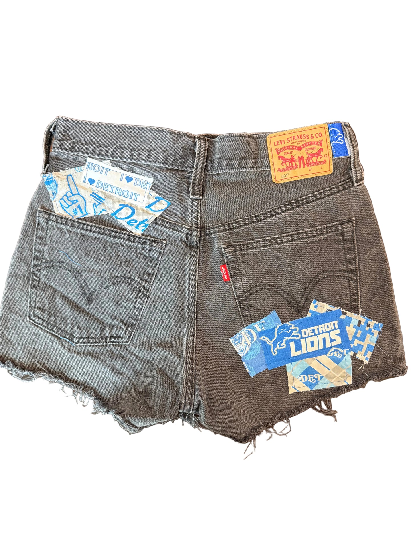 A pair of dark gray denim shorts with raw, frayed hems, featuring multiple colorful Levi's brand patches and labels attached to the back pockets, laid flat on a dark wooden surface.