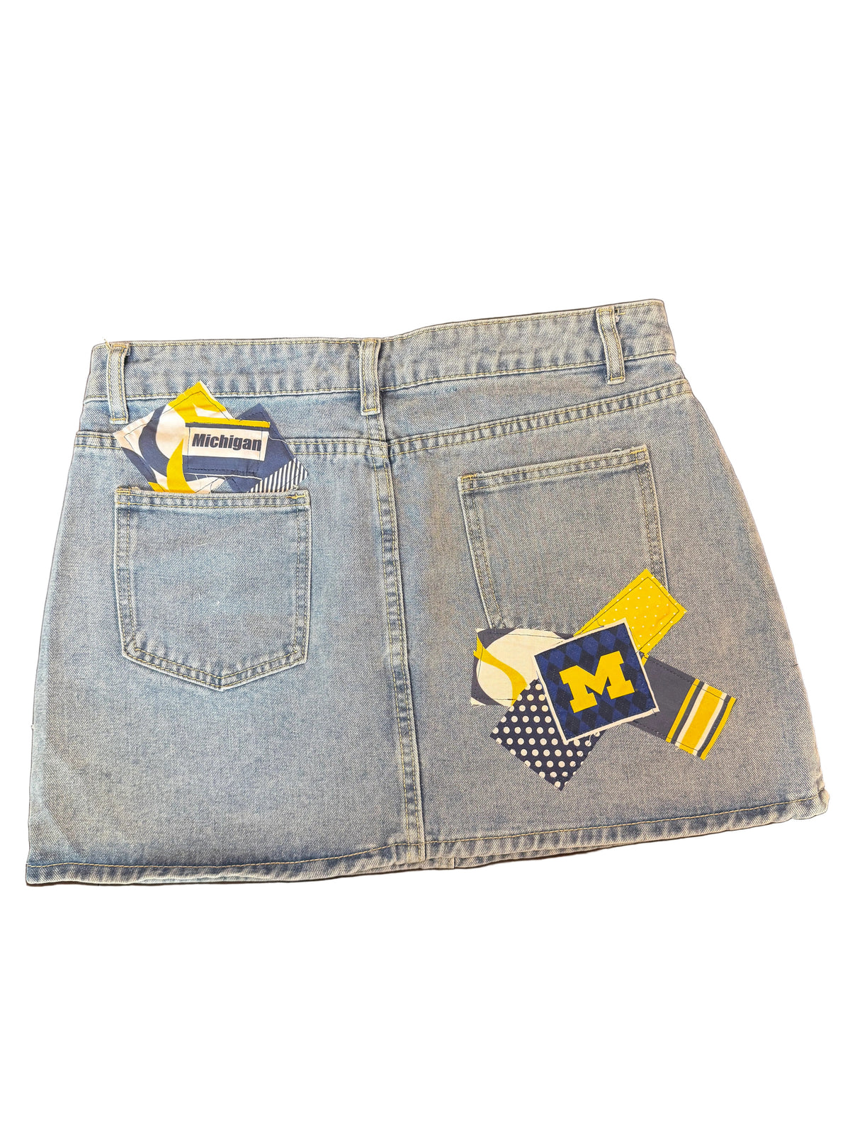 Light wash denim mini skirt with Michigan Wolverines themed patchwork back pocket designs, including a navy and yellow 'M' patch, a baseball patch, and striped details.