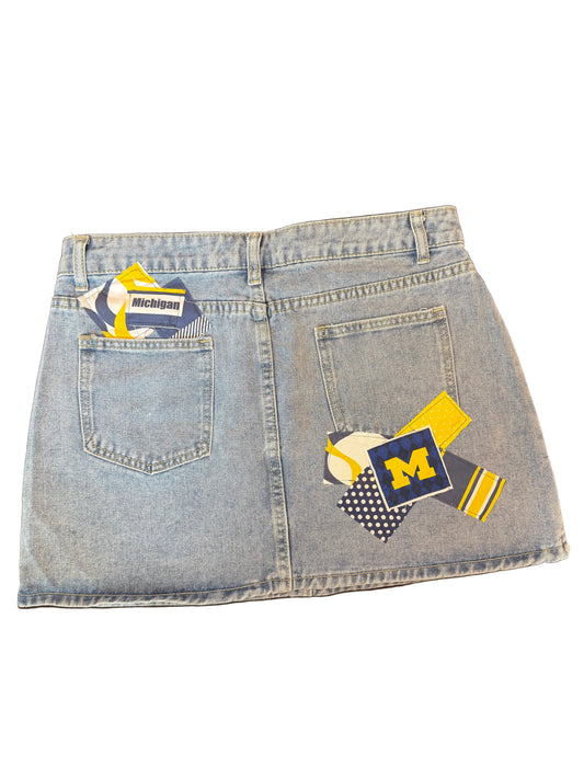 Light wash denim mini skirt with Michigan Wolverines themed patchwork back pocket designs, including a navy and yellow 'M' patch, a baseball patch, and striped details.
