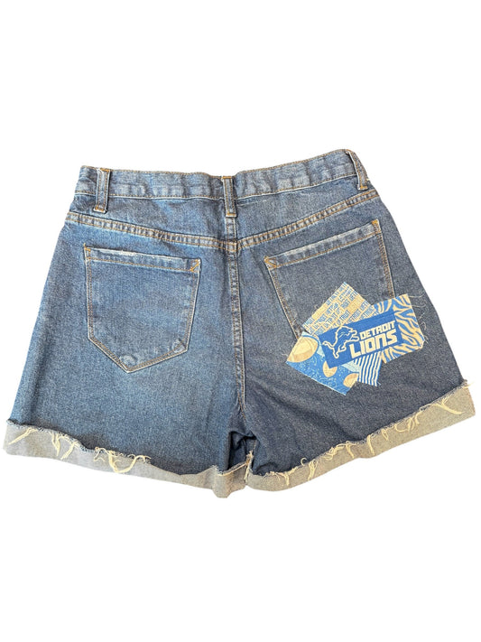 A pair of blue denim shorts with a unique asymmetrical design, featuring a large ripped section on one side with a beige lining and a blue graphic patch on the back pocket.