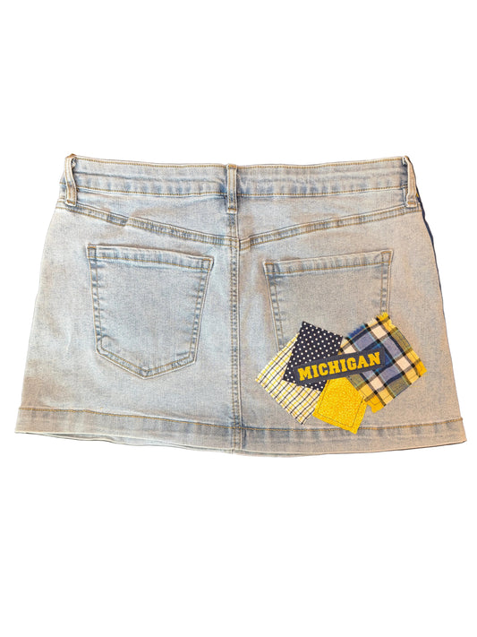 A light-wash denim mini skirt shown from the back, featuring two standard back pockets and a decorative patch on the right side with plaid fabrics and the word 'MICHIGAN' in yellow.