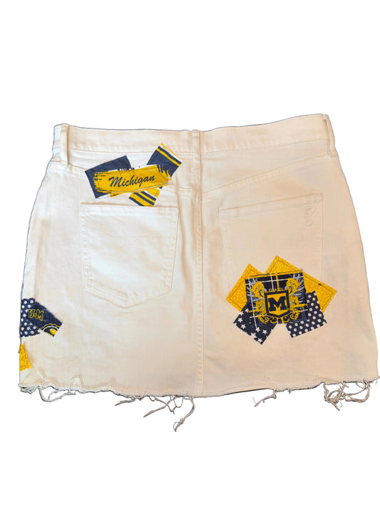 Off-white denim shorts with a raw, frayed hem and distressed details, featuring Michigan Wolverines themed patchwork fabric accents on the pockets and waistband.