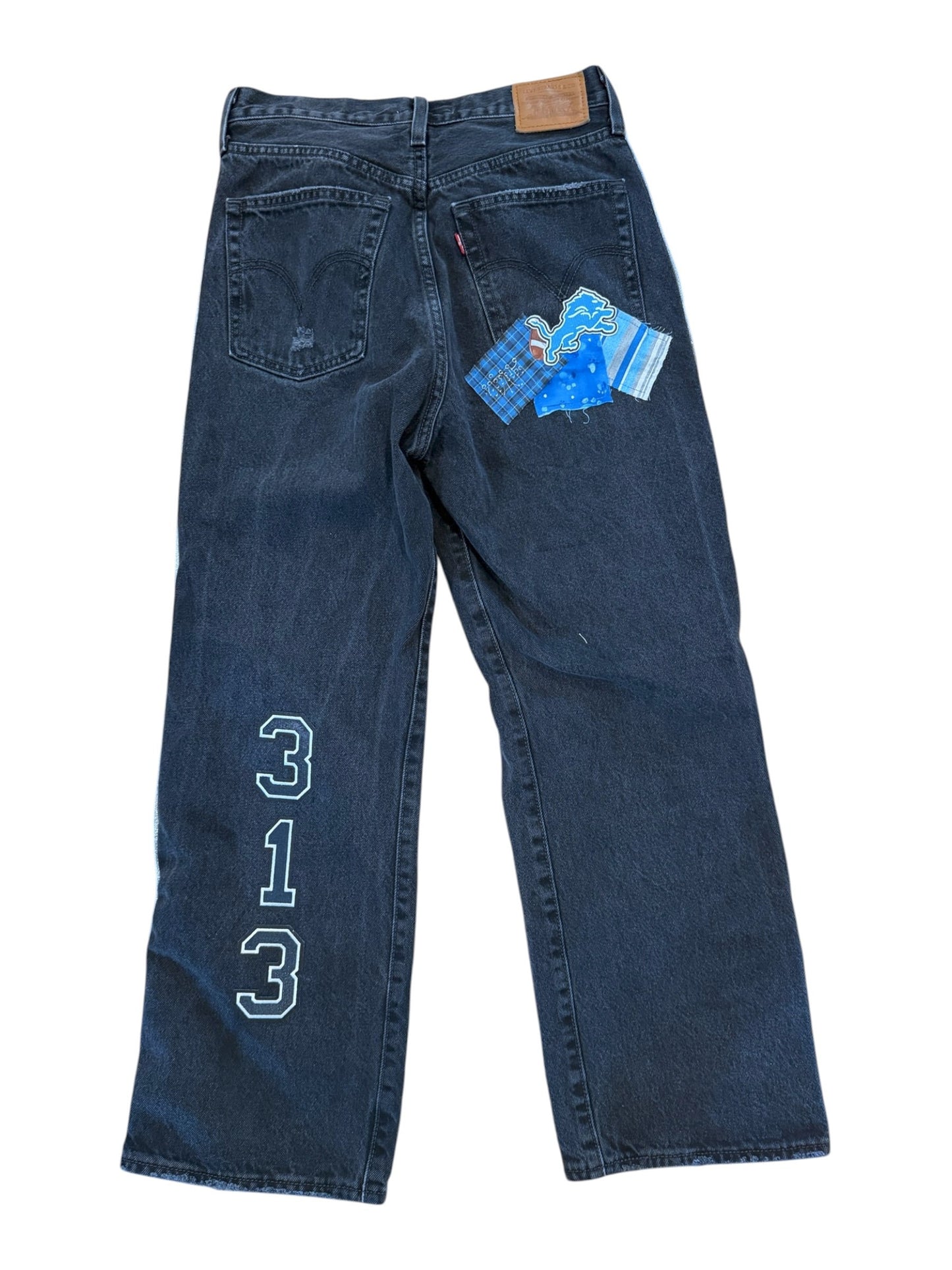 A pair of dark blue denim jeans laid flat on a white background, featuring a light blue embroidered geometric logo on the upper left leg and a blue patterned paper tag attached near the back pocket.
