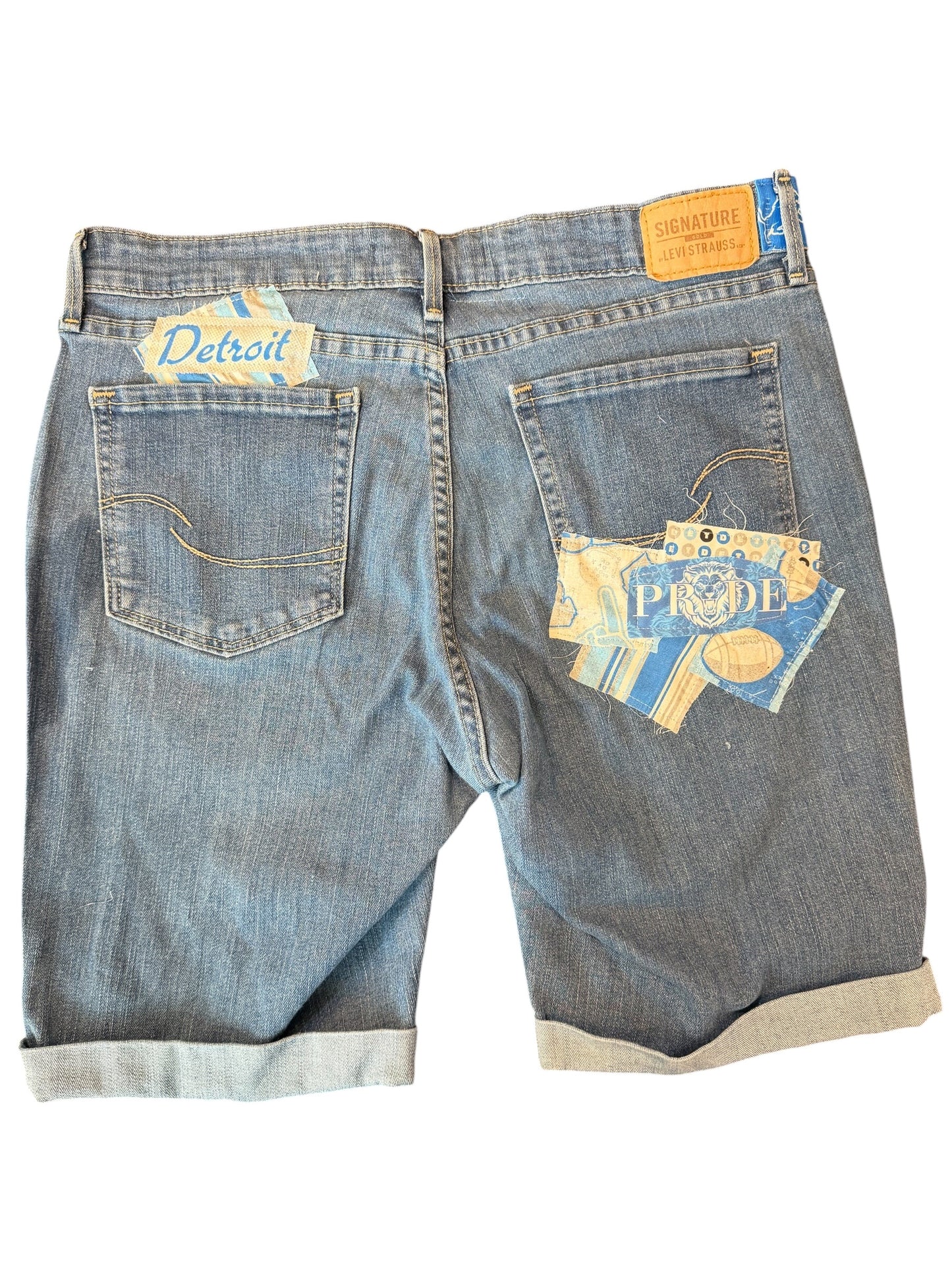 Back view of light blue denim shorts with rolled cuffs, featuring a 'Detroit' patch on the left back pocket and a larger 'PRIDE' patch on the right back pocket.