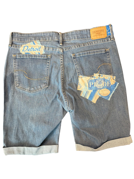 Back view of light blue denim shorts with rolled cuffs, featuring a 'Detroit' patch on the left back pocket and a larger 'PRIDE' patch on the right back pocket.