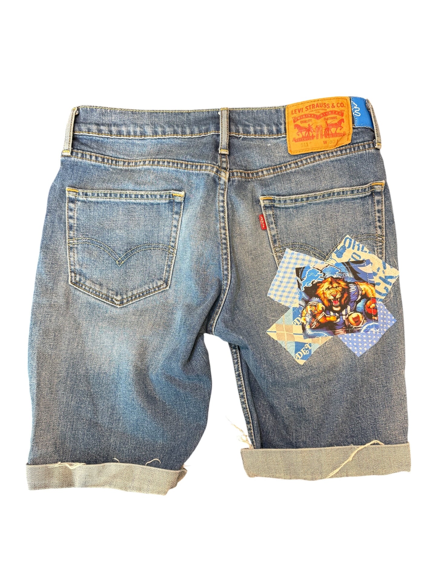 Back view of light blue denim shorts with rolled cuffs, featuring a colorful patchwork graphic on the right back pocket and a yellow Levi's waistband label.