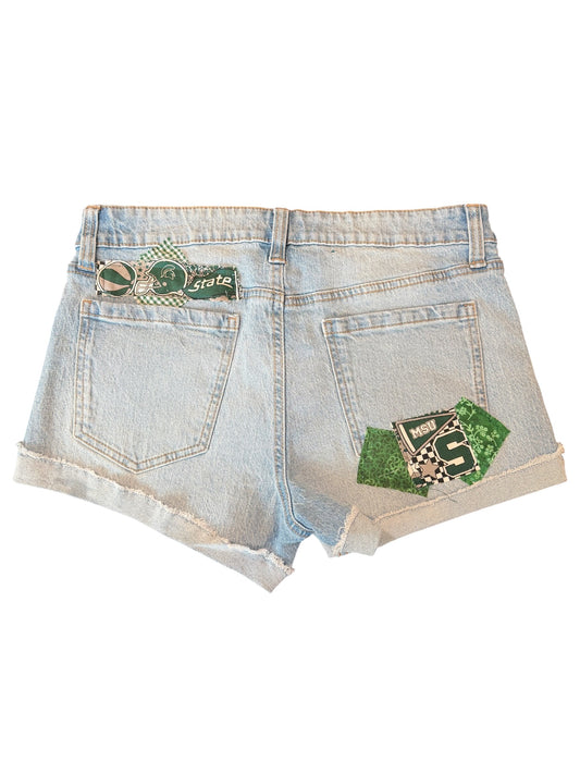 Light wash denim shorts with a distressed frayed hem, featuring a green and black patch on the back right pocket and a green and black letter 'S' patch on the back left pocket.
