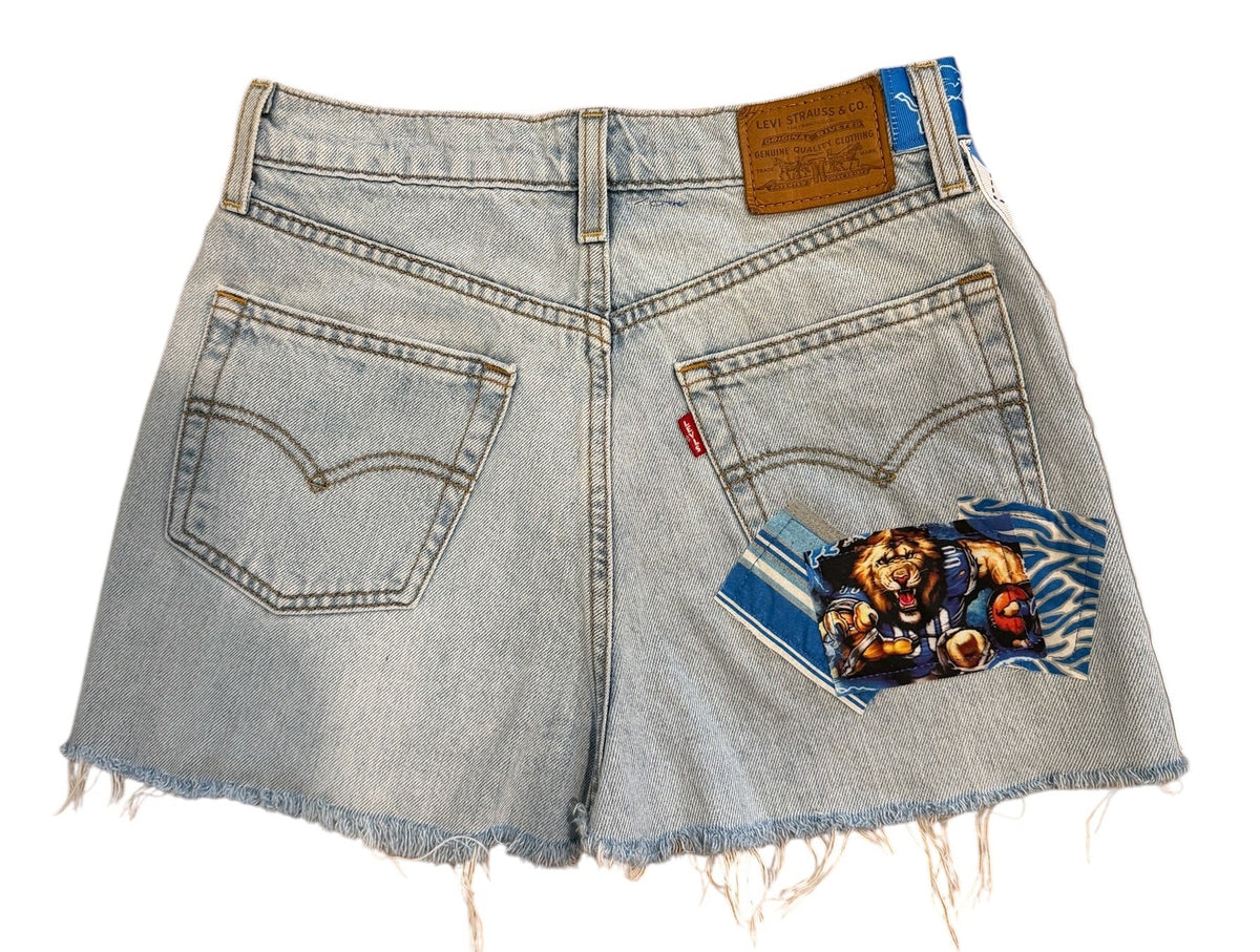 Light-wash denim shorts with a distressed, frayed hem and a colorful graphic patch on the back pocket, shown flat on a white background.