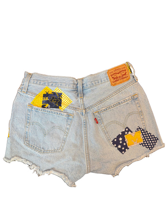 Light wash denim shorts with a raw frayed hem, featuring a yellow and black patch on the left back pocket, a yellow and black patch on the right back pocket, and a small orange Levi's brand patch on the waistband.