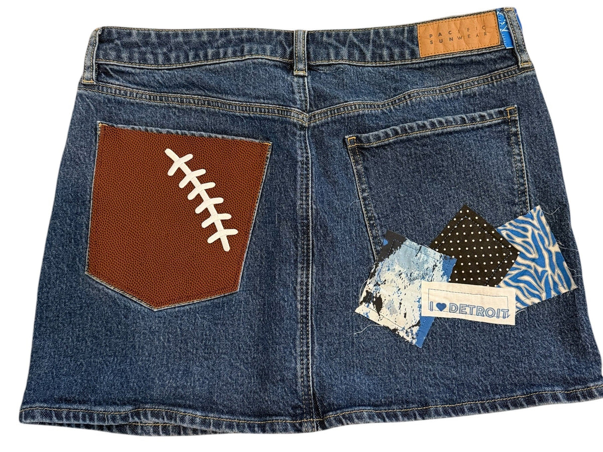 A pair of blue denim jeans laid flat on a white background, featuring a brown football-shaped patch on the back pocket with white lacing details, a patterned fabric patch on the front pocket, and a small tan brand label.