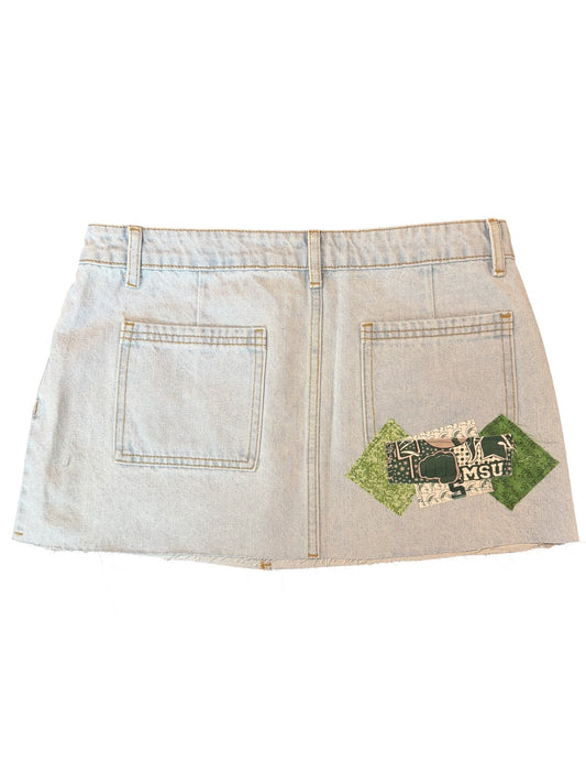 A light-wash denim apron or bib-style garment laid flat on a white background, featuring two front pockets and a green diamond-shaped patch with black text and graphic details on the lower left pocket area.
