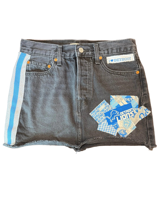 A pair of dark gray denim shorts with a blue and white patterned patch on the left leg and a light blue and white stripe along the hem, laid flat on a dark wooden table.