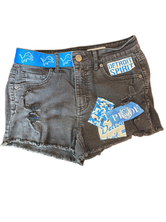 A pair of gray washed denim shorts with distressed details and raw frayed hems, laid flat on a dark wooden surface. The shorts feature a white and blue 'This is Hollister' patch on the front pocket and several blue and white brand tags attached.