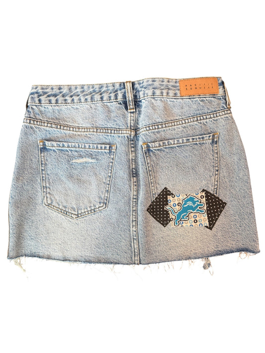 Light wash denim mini skirt shown from the back, featuring a distressed frayed hem, two back pockets, and a Detroit Lions patch on the right pocket.