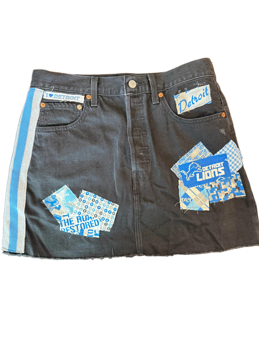A dark gray denim mini skirt with Detroit Lions patches and a blue and white side stripe, laid flat on a wooden floor.