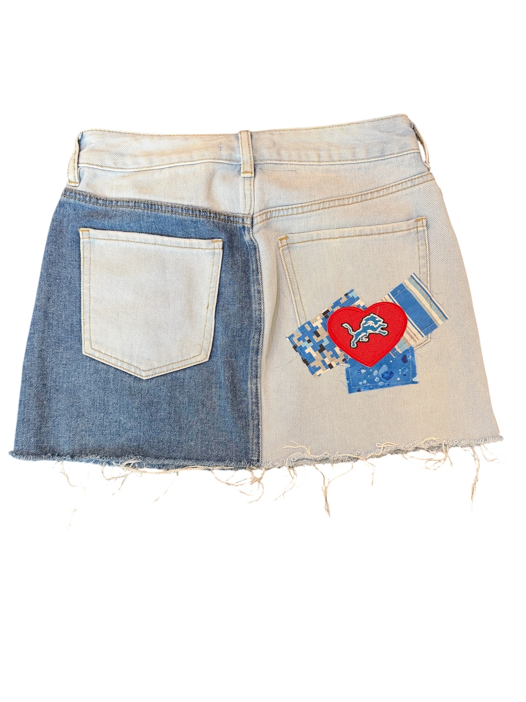 A denim mini skirt with a split design, featuring a medium blue denim left side and a light beige/cream right side. The back pocket on the right side is decorated with a red heart patch containing a white lion graphic and blue patterned fabric accents, while the left back pocket is plain.