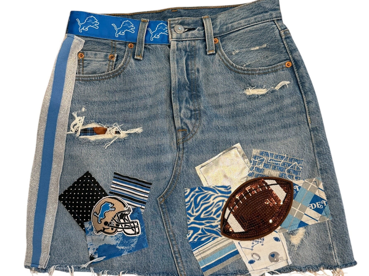A pair of light blue denim shorts featuring distressed rips, frayed edges, and various patchwork fabric and textile embellishments, including a football patch and patterned squares, laid flat on a white background.