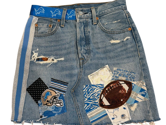 A pair of light blue denim shorts featuring distressed rips, frayed edges, and various patchwork fabric and textile embellishments, including a football patch and patterned squares, laid flat on a white background.
