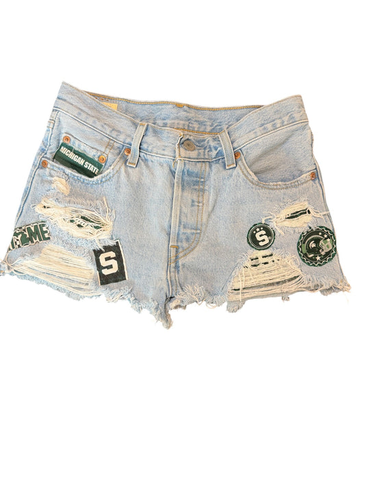 Light wash blue denim shorts with distressed rips, frayed hems, and multiple patches including a green 'Sesame Street' patch, a black patch with a white 'S', and circular green patches.