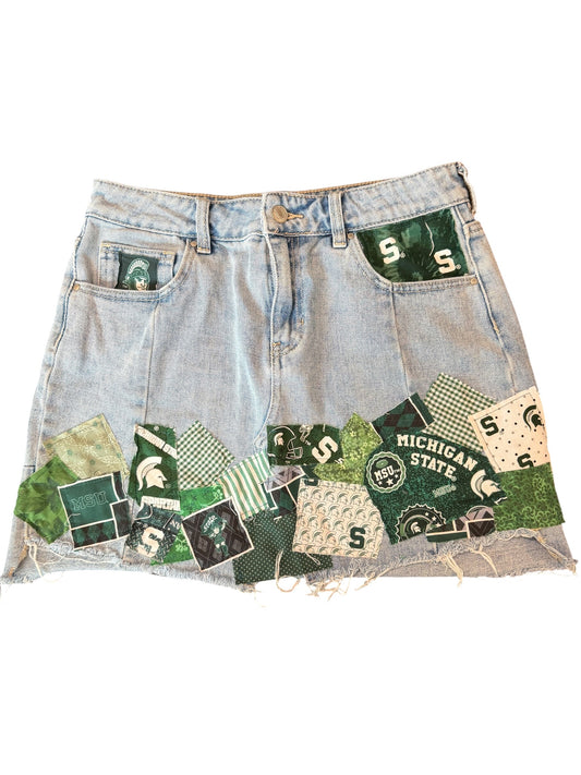 Light-wash denim shorts with a distressed frayed hem and a green Michigan State Spartans themed patchwork collage across the front pockets and lower hem.
