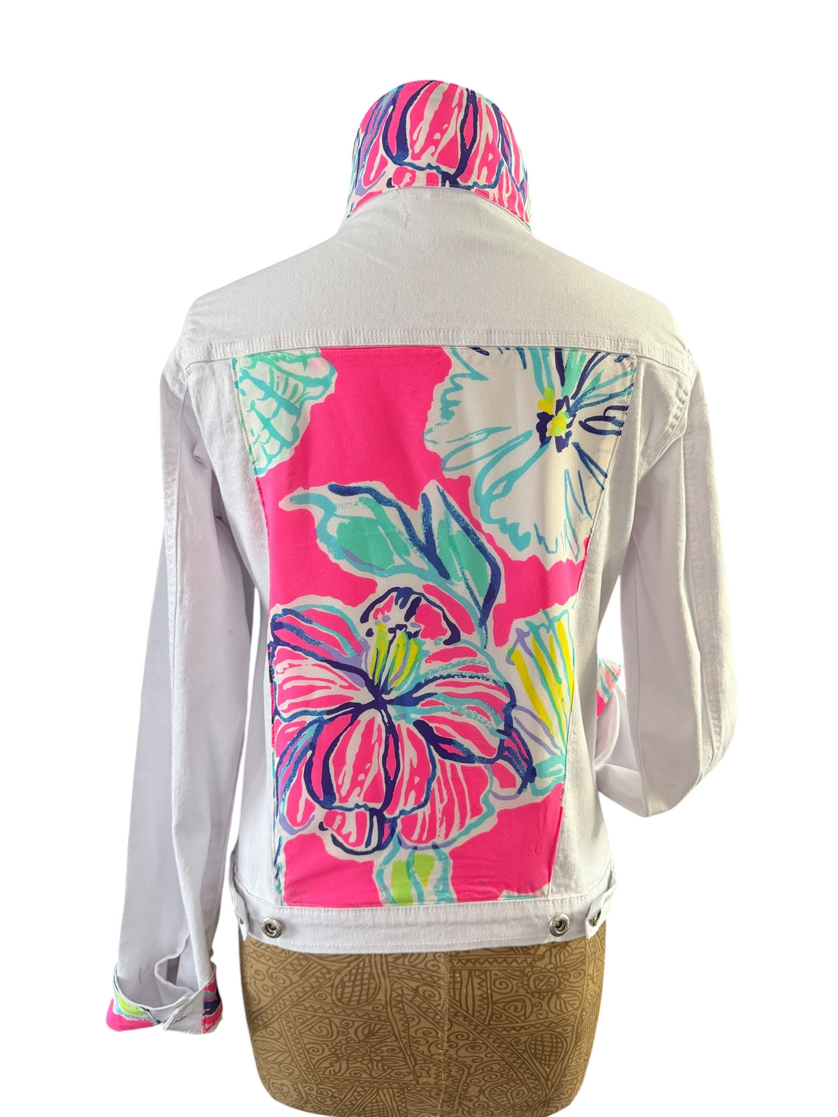 A white denim jacket shown from the back on a dress form, featuring a large vibrant pink tropical floral print panel on the back and a matching pink floral print hood, displayed against a white background.