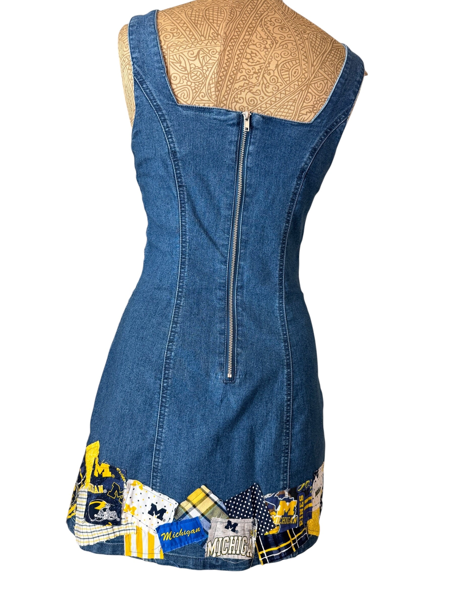 A sleeveless blue denim dress shown from the back on a mannequin torso, featuring a central zipper, a scoop neckline with a beige patterned insert, and a patchwork trim at the hem in blue, yellow, white, and black.