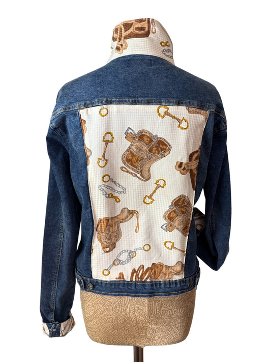 A denim jacket displayed on a dress form, showing the back view. The jacket features a solid blue denim body with a large cream-colored back panel decorated with brown horse-themed illustrations, including saddles, bridles, and horsebit details.