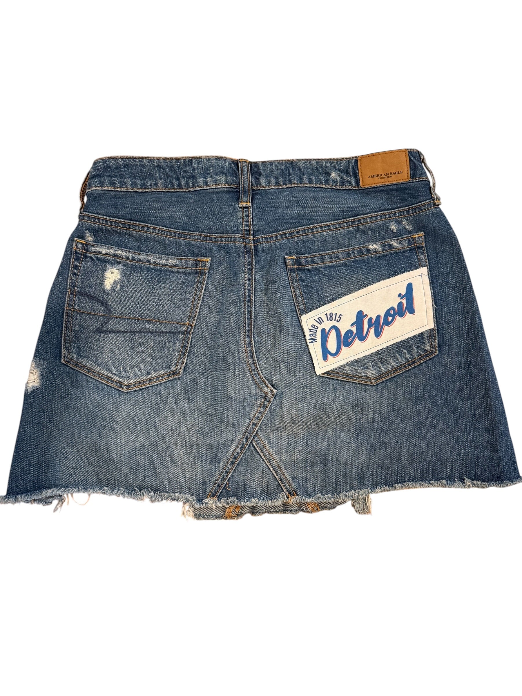 Back view of a distressed blue denim mini skirt with a frayed hem, featuring a 'Detroit' patch on the right pocket and a brown leather brand tab at the waistband.