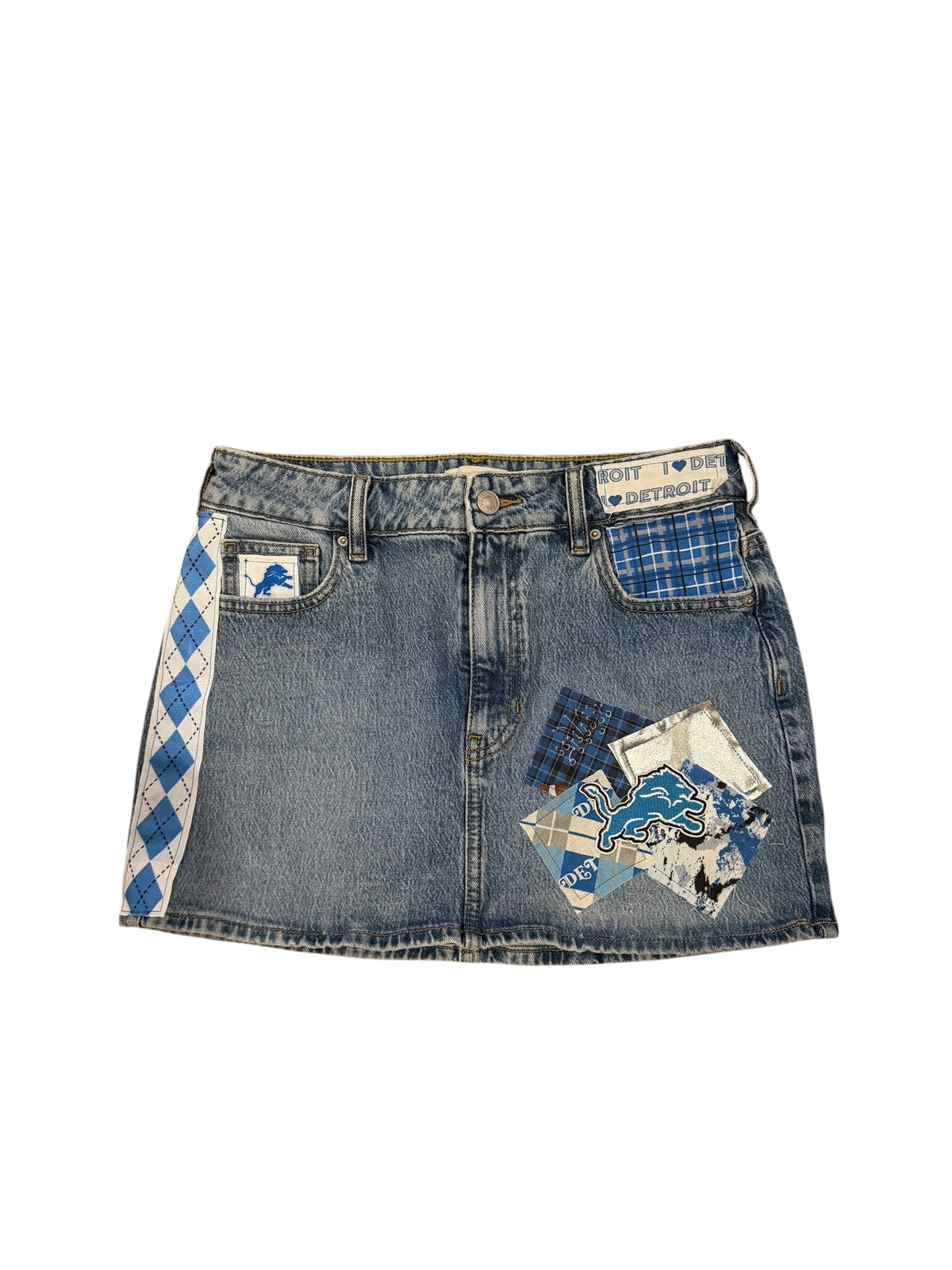 A denim mini skirt featuring a blue and white argyle pattern along the inner waistband, a blue and white plaid patch pocket, and a decorative collage of vintage-style postcards and fabric patches on the front left side.