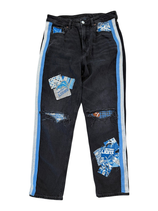 A pair of black distressed jeans with ripped details, blue and white graphic patches, and a light blue and white stripe down the outer side of each leg, shown on a plain white background.