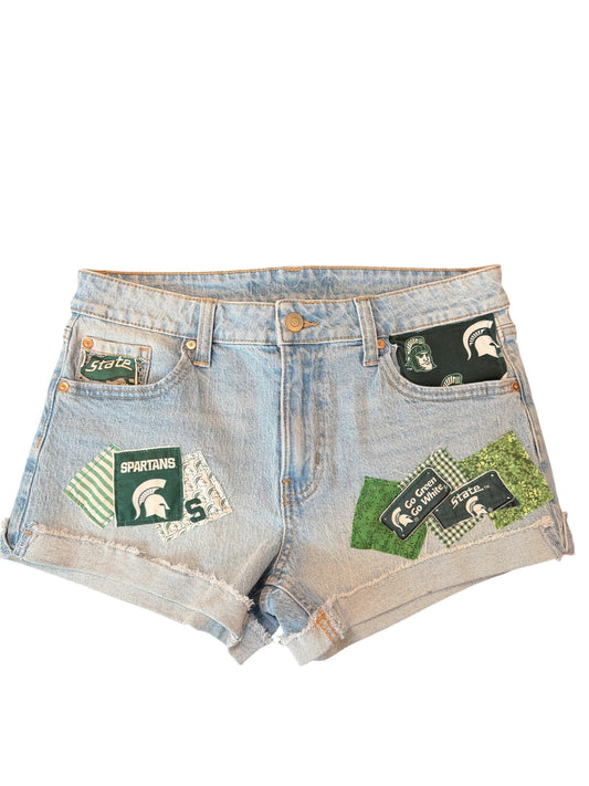 Light wash denim shorts with distressed frayed hems and multiple Michigan State Spartans themed patches sewn onto the front pockets and thighs.
