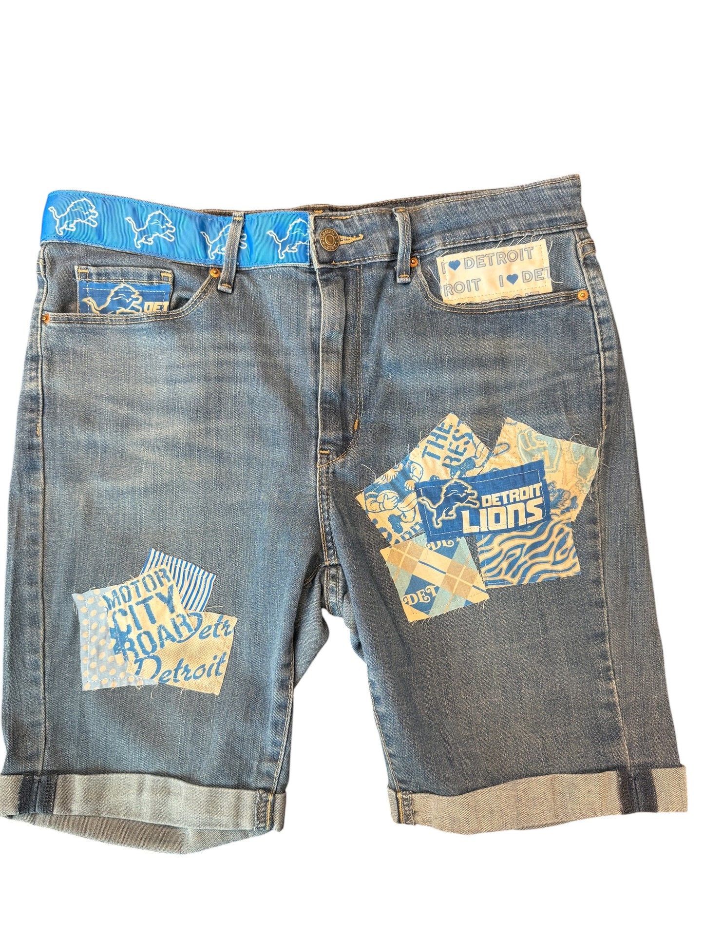 Light wash blue denim shorts with Detroit Lions patches on the front pockets and a blue patterned waistband, shown against a plain white background.