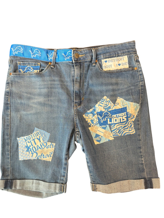 Light wash blue denim shorts with Detroit Lions patches on the front pockets and a blue patterned waistband, shown against a plain white background.