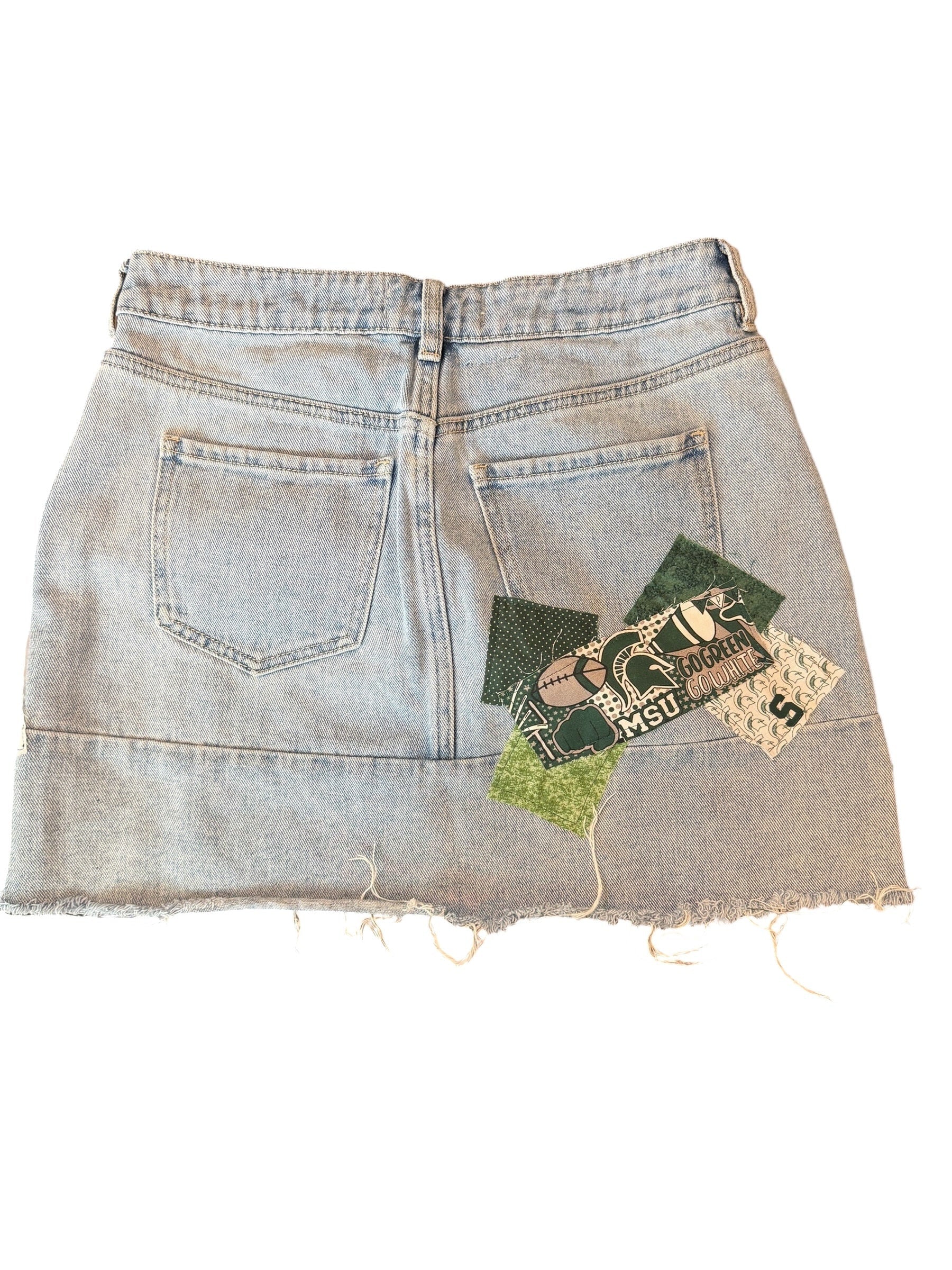 Light-wash denim shorts with a raw, frayed hem and decorative vintage-style fabric patches sewn onto the back pocket area, shown flat on a white background.