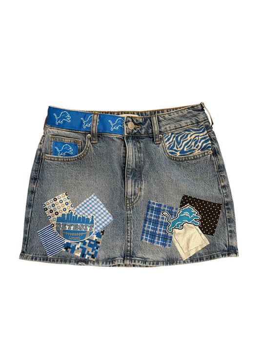 A light-wash denim mini skirt featuring Detroit Lions themed patchwork details, including a blue patterned waistband, a blue patterned pocket, and two large decorative patchwork panels on the front pockets with various blue, black, and beige fabrics.