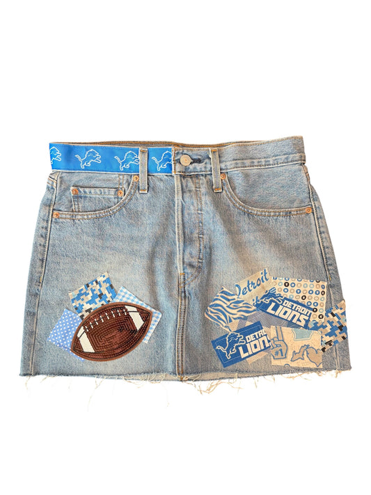 Light wash blue denim mini skirt with distressed frayed hems, featuring Detroit Lions themed patches including a football patch, a blue patterned patch, and a Michigan patch on the front.