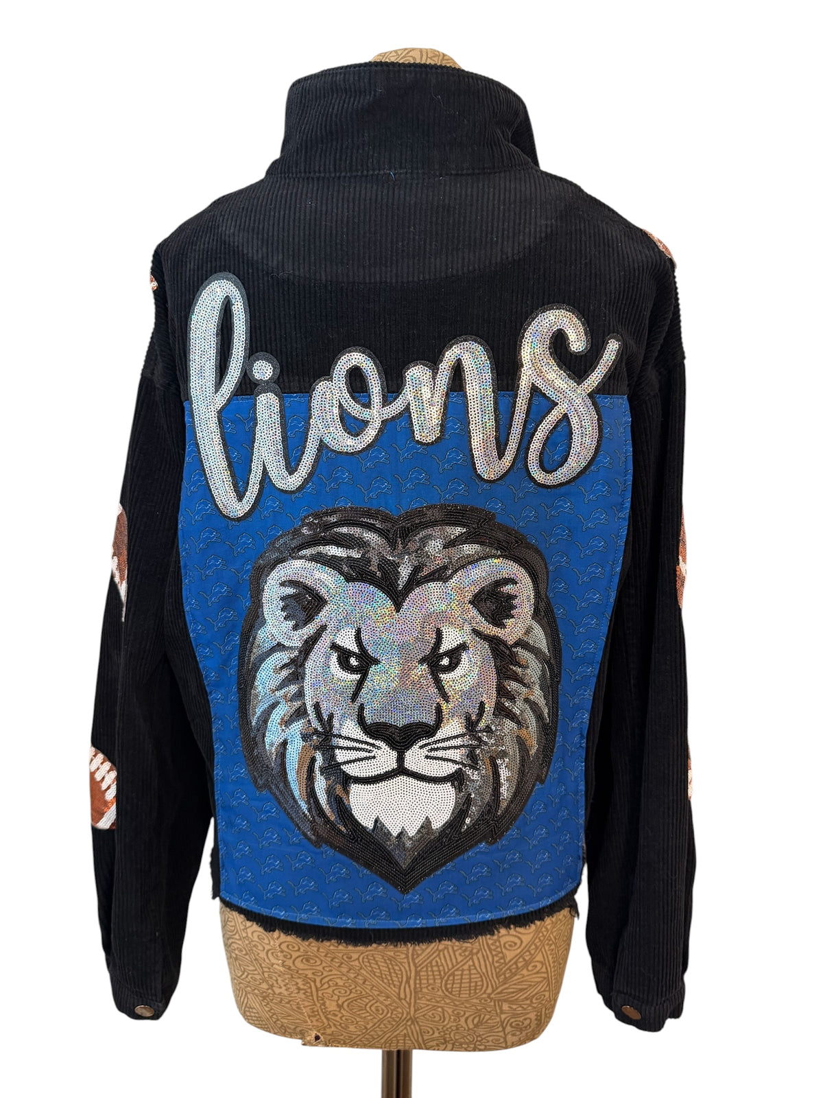 A black corduroy jacket shown from the back on a mannequin torso, featuring a large blue panel with a silver glittery 'lions' script and a detailed lion head graphic.