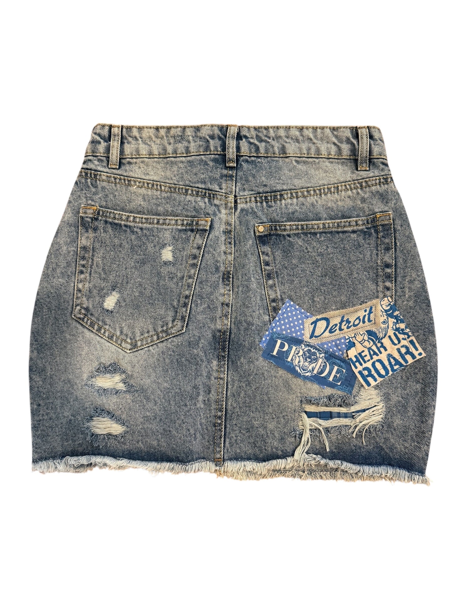 A distressed denim mini skirt with a frayed hem, featuring a blue and white 'Detroit Proud Teen Us Roar!' patch on the back right pocket, shown against a plain white background.