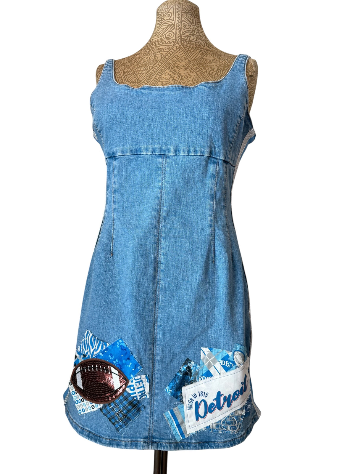 A light blue denim dress displayed on a tan mannequin torso against a white background. The dress features a scoop neckline, wide shoulder straps, and decorative printed patches on the lower front including a football graphic and a Detroit Tigers baseball card design.