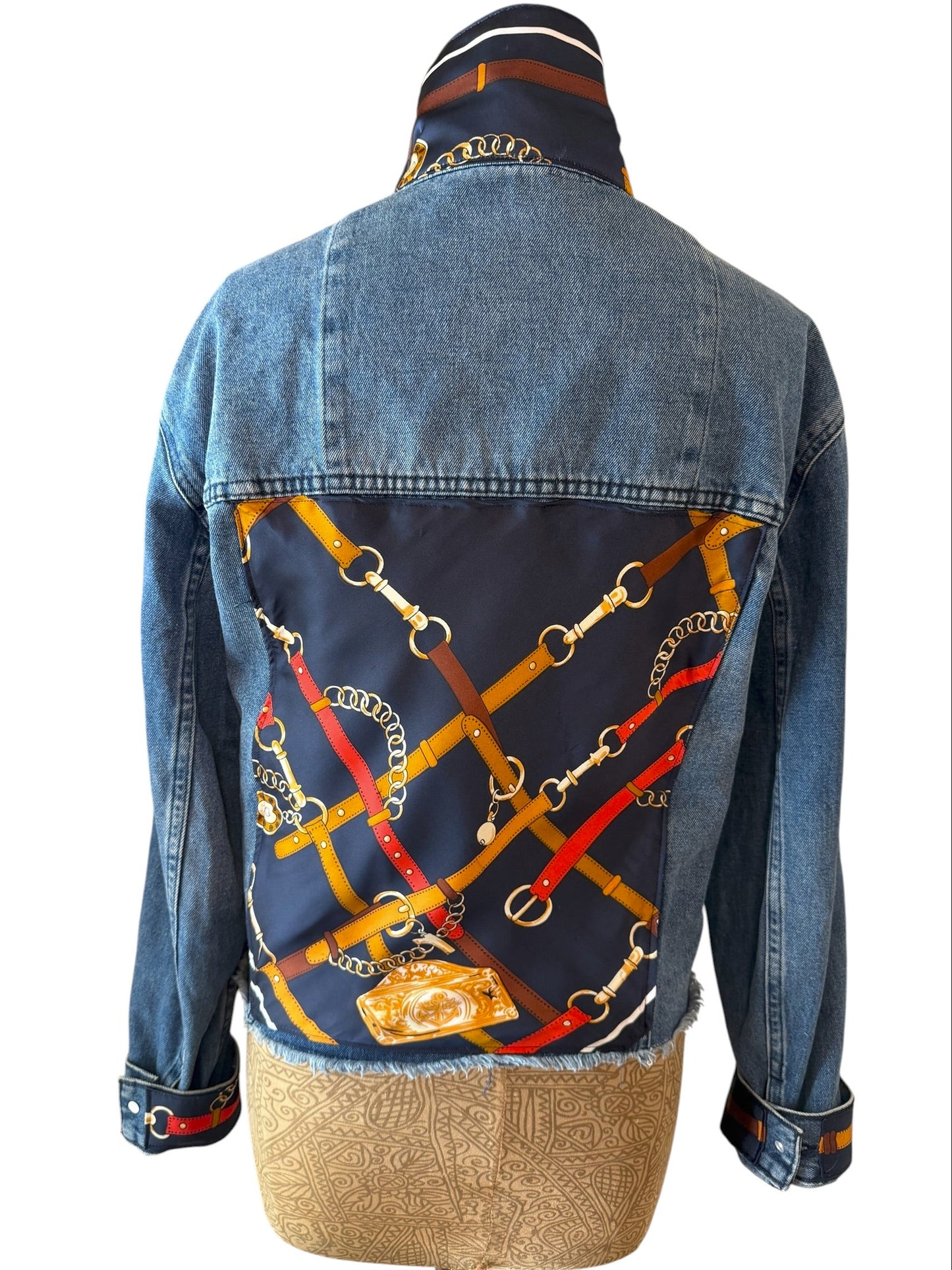 A denim jacket shown from the back on a dress form, featuring a large navy blue printed panel with gold horsebit chain graphics and red and orange accents.