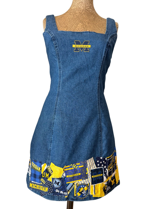 A blue denim pinafore dress displayed on a mannequin torso against a white background. The dress features a yellow and blue 'Michigan' logo patch on the chest and a colorful patchwork trim at the hem with 'Michigan' text, the 'M' logo, and sports-themed graphics.