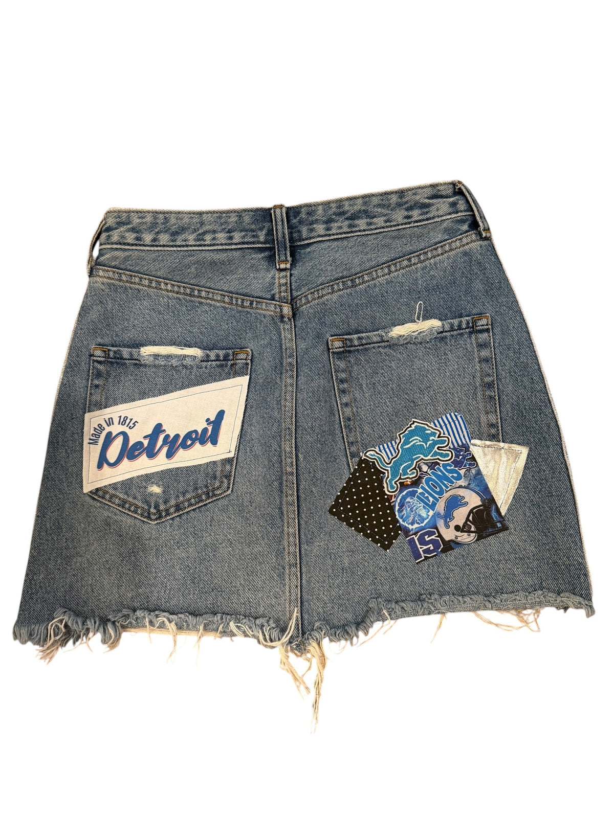 A pair of light blue denim shorts with raw, frayed edges, featuring a white 'Detroit' patch on the back pocket and a colorful graphic patch on the front pocket.