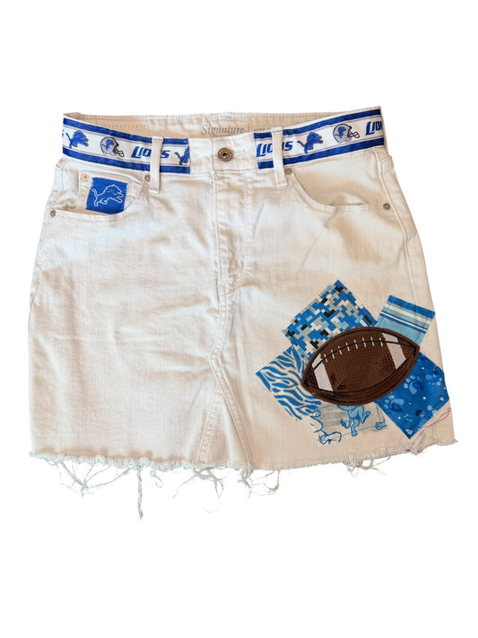 A pair of off-white denim shorts with a distressed frayed hem, featuring a blue and white Detroit Lions themed waistband, a blue patch on the pocket, and a blue-and-white patterned football patch on the front.