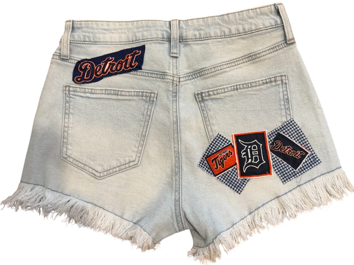 Light wash denim shorts with frayed hems, featuring decorative back pocket patches with 'Detroit' text and a gingham pattern, shown on a plain white background.