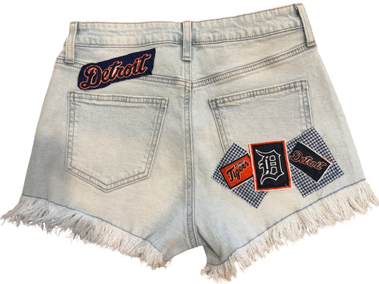 Light wash denim shorts with frayed hems, featuring decorative back pocket patches with 'Detroit' text and a gingham pattern, shown on a plain white background.